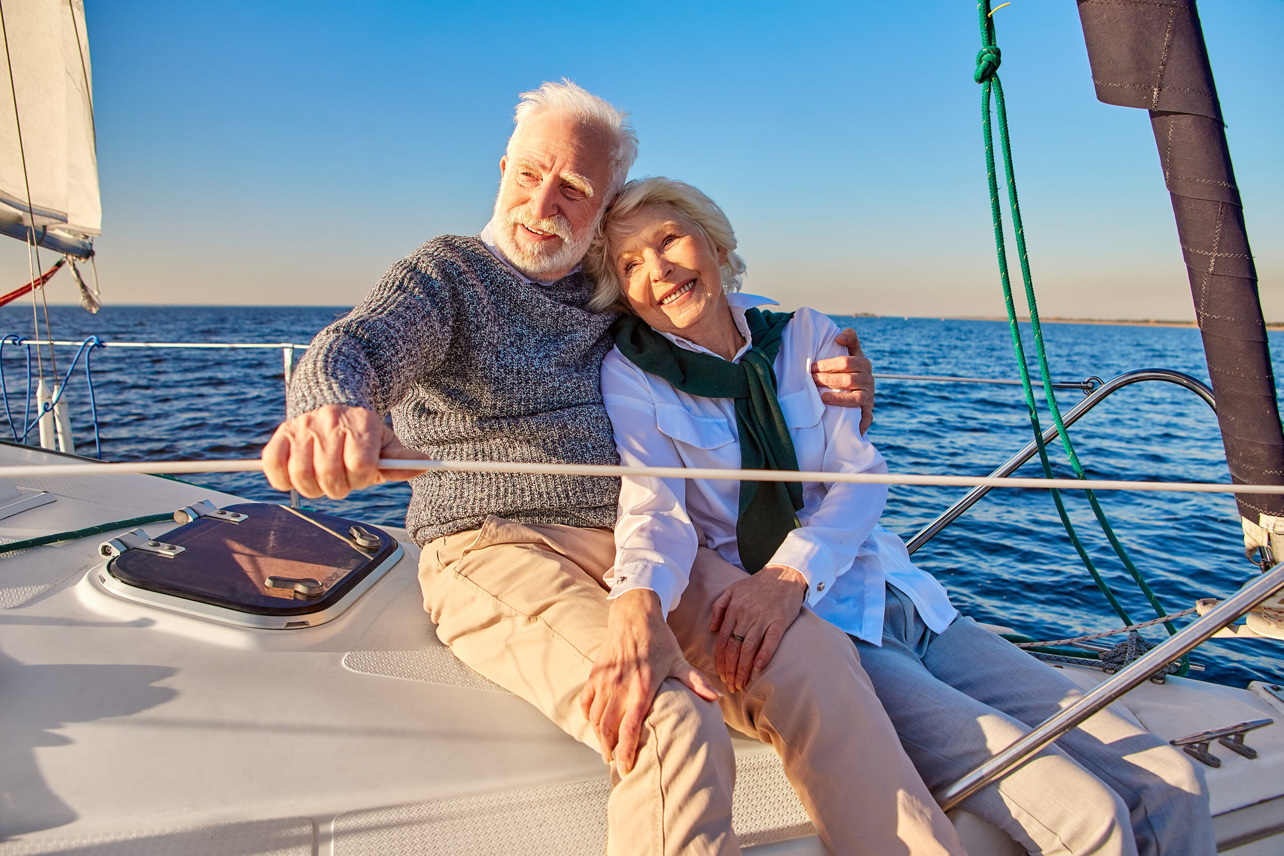 Happy beautiful senior family couple hugging and smiling while relaxing on a sail boat or yacht deck floating in a calm blue sea, enjoying amazing view