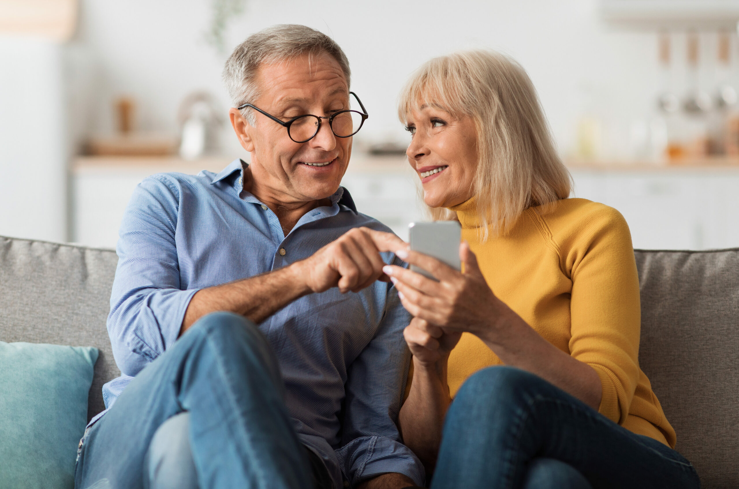 Senior Couple Texting Via Smartphone Sitting On Sofa At Home